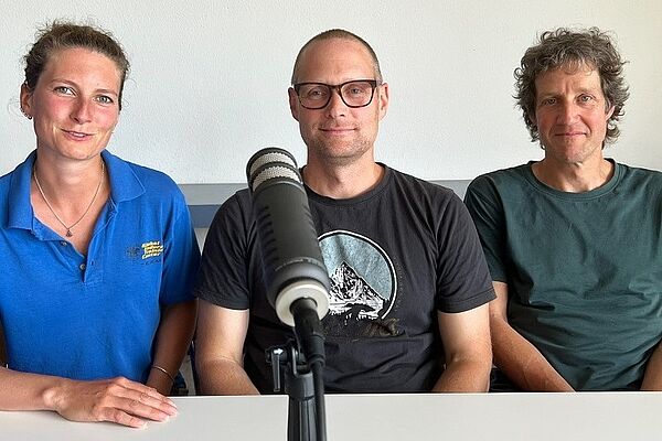 Die drei genannten Personen am Tisch sitzend vor dem Podcast-Mikrophon