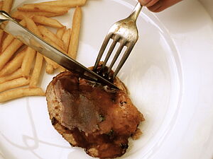 Ein Teller mit Pommes und einem Stück Fleisch, Gabel und Messer schneiden das Fleisch.