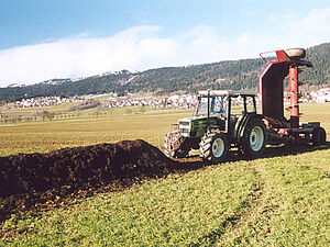 Mistkomposthaufen auf einem Acker, nebenbei ist ein Traktor und eine Kompostwendemaschine zu sehen.