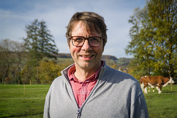 Ein Mann auf einer Wiese mit einer Kuh im Hintergrund.