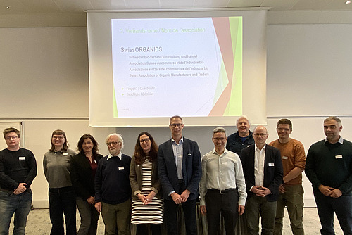 Eine Gruppe von 11 Menschen posiert vor einer Powerpoint-Präsentation.