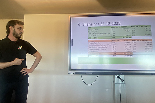 Ein Mann mit Bart und Brille steht neben einem Bildschirm mit einer Powerpoint-Präsentation.