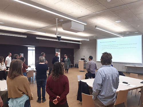 Einige Menschen stehen unregelmässig verteilt in einem Saal mit einer Powerpoint-Präsentation.
