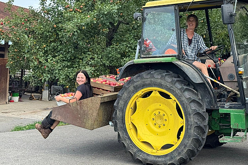 Eine Frau sitzt auf dem Fahrersitz eines grünen Traktors, eine weitere Frau sitzt hinten auf einer Traktorschaufel mit Äpfeln. 