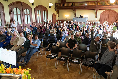 Mitglieder des Vereins für biologisch-dynamische Landwirtschaft sitzen in einem Saal und halten ihre Stimmkarten nach oben.