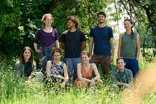 Ein Gruppe von Menschen unterschiedlichen Alters im Gras und im Schatten einiger Bäume.