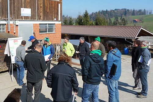 zehn Männer und zwei Frauen stehen im Kreis vor einer Scheune mit Posterständer