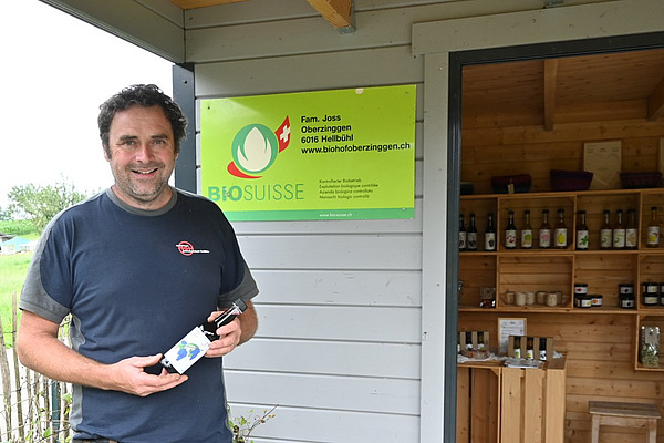 Ein Mann steht mit einer Flasche in der Hand vor einem Hofladen. 