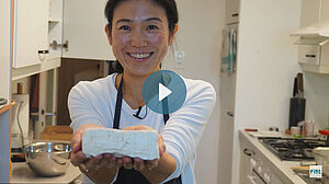 Tofu-Meisterin Mariko Kaufmann präsentiert einen frisch hergestellten Tofuwürfel; Bild inklusive Filmstart-Button.