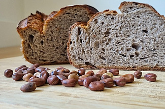 Auf einem Tisch steht ein angeschnittener Laib Brot, davor liegen Ackerbohnensamen. 