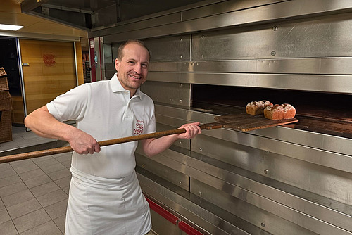 Ein weiss gekleideter Mann mit weissem T-Shirt und Schürze vor einem Backofen. 
