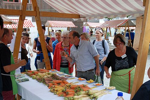 Eine Verkäuferin und ein Verkäufer bieten der Kundschaft an einem Marktstand verschiedene Karottensorten an.