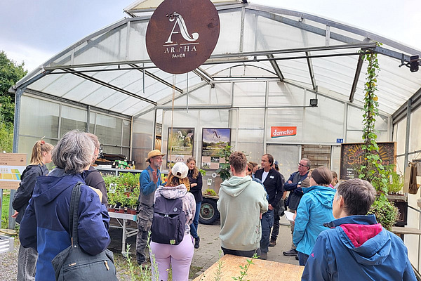 Eine Gruppe von Menschne mit dem Rücken zur Kamera, dahinter ein Treibhaus, davor ein Referent.