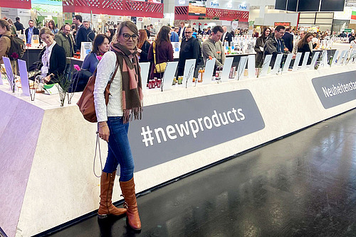 Eine Frau steht vor einem langgezogenen Korpus an einem Messestand, bestückt mit Produkten, im Hintergrund viele Menschen. 
