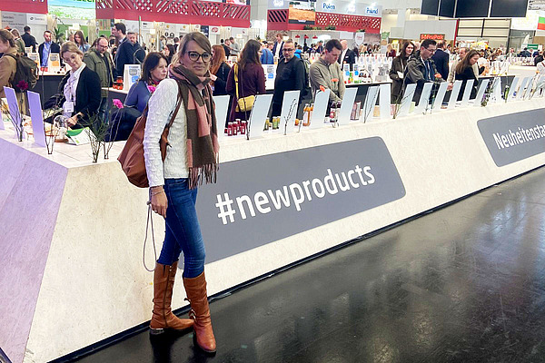 Eine Frau steht vor einem langgezogenen Korpus an einem Messestand, bestückt mit Produkten, im Hintergrund viele Menschen. 