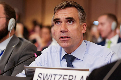 Ein Mann in einem Saal mit Tischmikrofon und Schild mit Switzerland drauf.