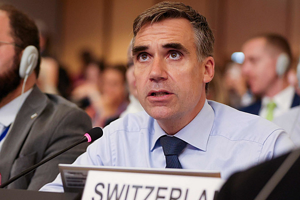 Ein Mann in einem Saal mit Tischmikrofon und Schild mit Switzerland drauf.