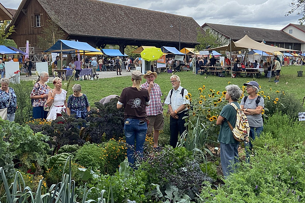 Menschen in einem Gemüsegarten, dahinter Marktstände. 