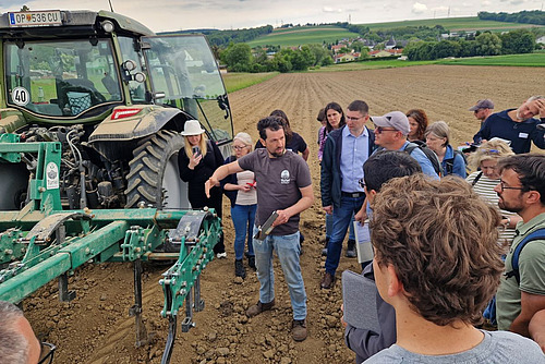 Landwirt steht neben einem Traktor auf dem Acker und spricht zu einer Gruppe.