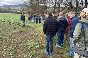 Eine Gruppe Personen steht auf einem Feld im Winter, eine Person erkärt etwas.