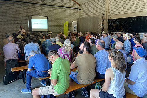 Ein gut gefüllter Saal mit einem Referenten im Hintergrund. 