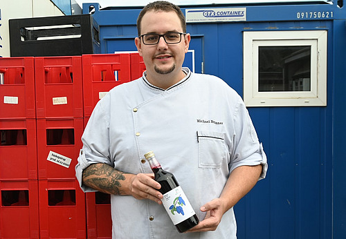 Ein Mann hält vor einer Beige Harassen und einem Bürocontainer eine Flasche. 