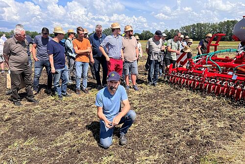 Ein Mann hockt auf einem Feld und erklärt etwas zu dem Boden. Im Hintergrund stehen die Zuhörer*innen.