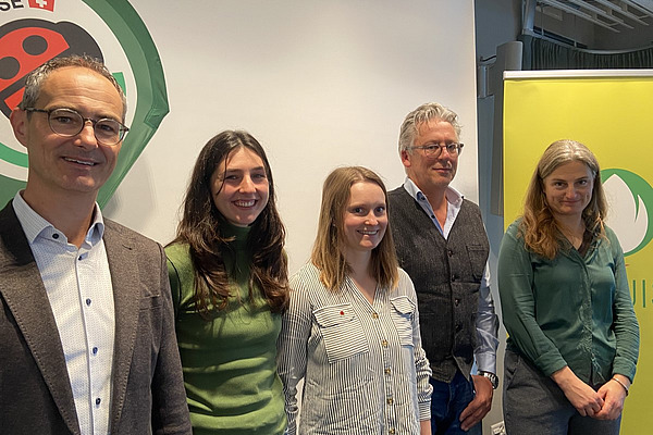 Ein Gruppenbild mit zwei Männern und drei Frauen vor einer Wand mit zwei Logos.