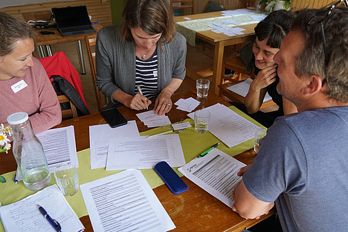 4 Personen sitzen an einem Tisch über Papiere gebeugt, eine Person schreibt etwas. 