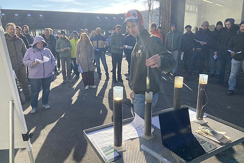 Eine Gruppe von Menschen beobachtet in der Wintersonne eine Demonstration einer Güllegehaltsmessung mit mehreren Messbechern. 