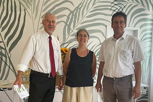 Die drei Personen posieren vor einer mit Palmblättern bemalten Wand.