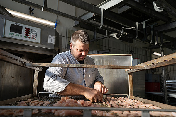 Zu sehen ist ein Metzger in Arbeitskleidung, der seine Tätigkeit mit Sorgfalt ausübt. Im Vordergrund sind Streifen eines Tierprodukts zu erkennen.