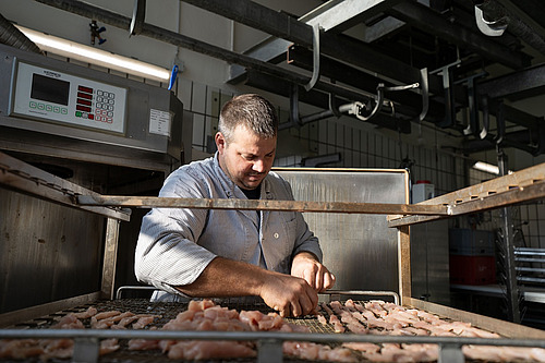 Zu sehen ist ein Metzger in Arbeitskleidung, der seine Tätigkeit mit Sorgfalt ausübt. Im Vordergrund sind Streifen eines Tierprodukts zu erkennen.