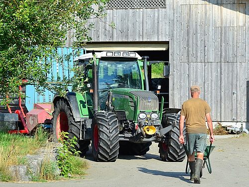 Ein Bauer läuft mit einer Schubkarre und dem Rücken zur Kamera Richtung Traktor und Scheune