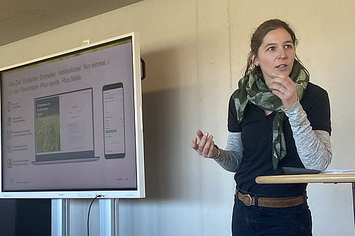 Eine Frau referiert neben einem Bildschirm mit einer Powerpoint-Präsentation. 