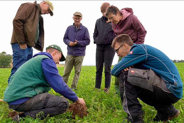 Eine Menschengruppe steht im Grünland rund um einen mit Erde gefüllte Spaten. 