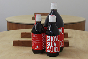 Auf einem Tisch stehen drei dunkle Flaschen mit rotem Etikett und der Aufschrift Shoyu Soja Sauce.  