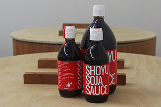 Auf einem Tisch stehen drei dunkle Flaschen in verschiedenen Grössen mit einem roten Etikett und der Aufschrift « Shoyu Soja Sauce ».  