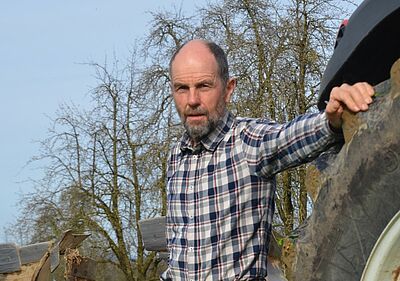 Ein Mann lehnt an einem Traktorreifen und schaut in die Kamera. 