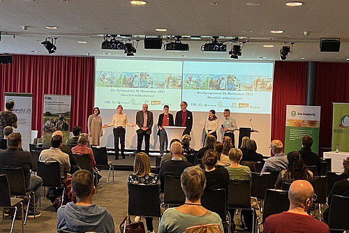 Ein Saal mit sitzenden Zuschauerinnen und Zuschauern sowie einer doppelten Powerpoint-Präsentation und sieben Leuten auf der Bühne.
