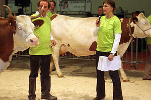 Anet Spengler steht vor einer Kuh , sie spricht ins Mikrofon und gibt ein Kommentar zu dieser Kuh. Hinter der Kuh ist Biolandwirt Andreas Wüthrich zu sehen. Das Bild stammt von einer Viehschau in Lausanne im Januar 2012.