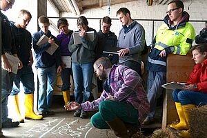 Eine Gruppe junger Personen steht im Kreis in einem Stall um einen Dozenten, der mit Kreide eine Formel auf den Boden schreibt. 