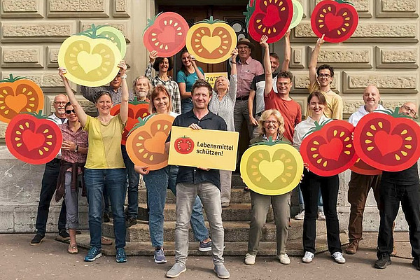 Eine Gruppe von Menschen posiert vor einer Steinmauer mit Schildern, worauf stilisierte Tomaten abgebildet sind..