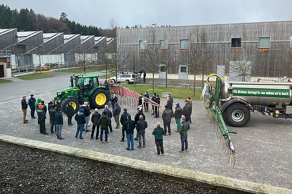 Menschen stehen auf einem Platz und betrachten einen Traktor mit Schleppschlauch und ein Güllefass.
