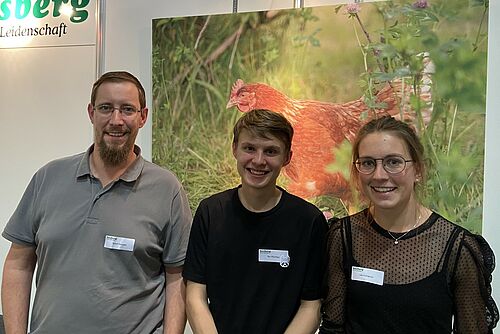 Drei Personen lächeln in die Kamera. Im Hintergrund ist ein Bild von einem Huhn im Gras. 