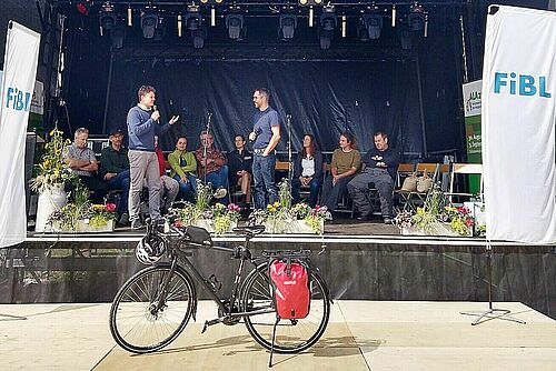 Im Hintergrund stehen mehrere Personen auf einer Bühne, rechts eine FiBL-Flagge im Vordergrund steht ein Velo