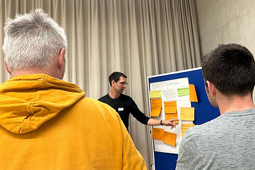 Mann steht vor einer Pinnwand und erklärt den Zuhörenden was auf den Post-its steht.