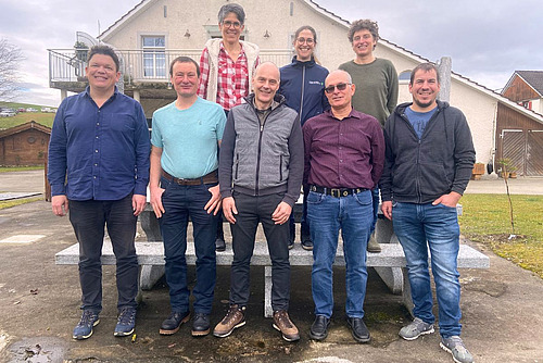 Gruppenbild mit zwei Reihen, davon eine leicht erhöht, vor einer ländlichen Gebäuefassade.