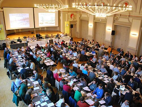 Die Delegierten an der Arbeit im Saal mit zwei Kronleuchten und zwei bebeamten Leinwänden