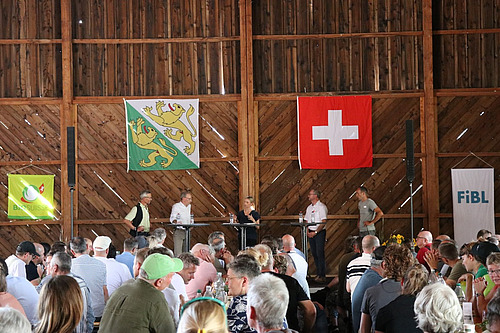 Podium in einem gut gefüllten Saal, auf der Bühne vier Männer und eine Frau in der Mitte als Leiterin.